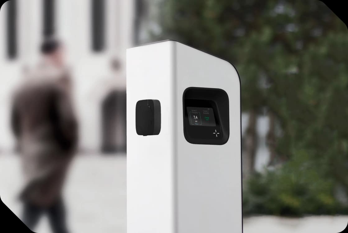 A WattsWhere charging pillar on a city street, a pedestrian passing in the blurred background